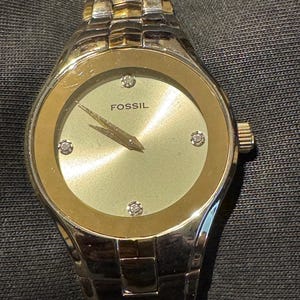 May include: A Fossil watch with a silver and gold-tone metal band and a round face. The watch face is light green with gold-tone accents and four small diamond-like markers. The word "FOSSIL" is printed on the watch face.