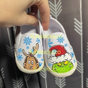 May include: A pair of white baby shoes with a Christmas theme. One shoe features a Grinch wearing a Santa hat and the other shoe features the Grinch as a reindeer. Both shoes have blue snowflakes on them.