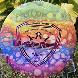 May include: A multi-coloured Dynamic Discs Maverick disc golf disc. The disc has a swirling pattern of rainbow hues. Text on the disc reads "DYNAMIC DISCS" and "MAVERICK". The disc is an understable fairway driver.