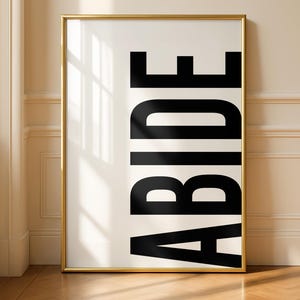 Op de afbeelding: Een ingelijste kunstdruk met het woord "ABIDE" in vette, zwarte letters op een crèmekleurige achtergrond. De gouden lijst contrasteert met de neutrale tinten van de kamer, die een houten vloer en een raam heeft.