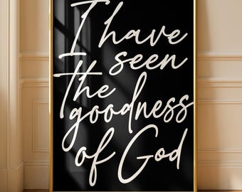 May include: A framed art print with the text "I have seen the goodness of God" in white script on a black background. The frame is gold, and the print is displayed against a neutral wall and wooden floor.