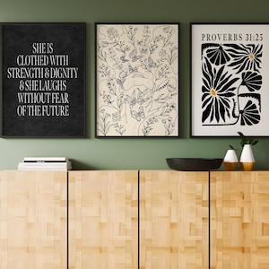 Könnte beinhalten: Drei schwarze gerahmte Drucke mit weißen Hintergründen und schwarzem Text und Illustrationen. Der linke Druck hat den Text "She is clothed with strength & dignity & she laughs without fear of the future". Der mittlere Druck hat eine Schwarzweißillustration von Blumen und Schmetterlingen. Der rechte Druck hat den Text "Proverbs 31:25" und eine Schwarzweißillustration von stilisierten Blumen.