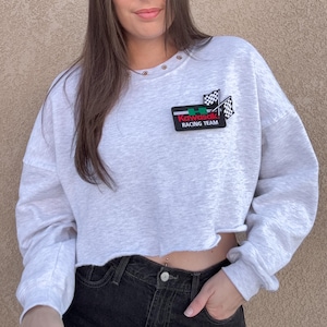 May include: A light gray cropped sweatshirt with long sleeves. The sweatshirt features a Kawasaki Racing Team patch with a checkered flag design. The raw hem and crew neck add a casual touch to this athletic-inspired top.