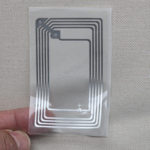 Puede incluir: Una pegatina rectangular transparente con un diseño plateado similar a un circuito. La pegatina tiene una serie de líneas paralelas que forman una forma rectangular con esquinas redondeadas. Un signo más es visible en la parte inferior. La pegatina está sujeta por una mano.