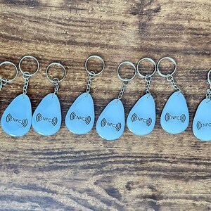 May include: A group of light blue NFC keyrings with silver rings, displayed on a wooden surface. Each keyring has a teardrop shape and features the text "NFC" with a radio wave symbol.
