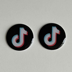 May include: Two black round buttons featuring the TikTok logo. The logo is a white musical note with a red and blue outline. The buttons are glossy and appear to be made of plastic or metal.