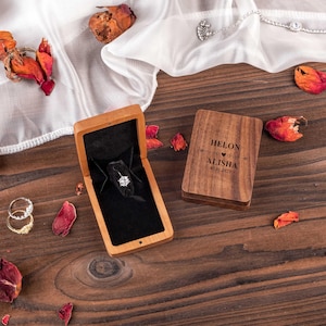 Puede incluir: Una caja de anillo de madera con un interior de terciopelo negro. La caja está grabada con los nombres "Helon & Alisha" y la fecha "02.01.2023". Un anillo de diamantes está dentro de la caja.