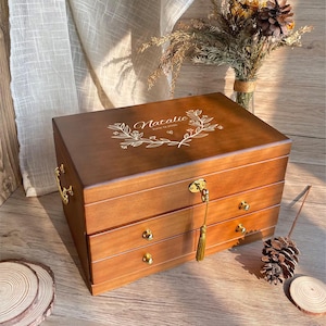 以下が含まれることがあります： 3つの引き出しと金色の金具が付いた木製のジュエリーボックス。「Natalie」という名前が装飾的なフォントで刻まれ、花の要素で囲まれています。鍵がロックからぶら下がっています。