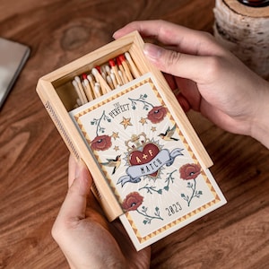 Op de afbeelding: Een houten luciferdoosje met een decoratieve kaart met een hart, kroon en bloemmotief. De kaart leest "The Perfect Match" en "A+F Match" met het jaar "2025". De luciferkoppen zijn rood.
