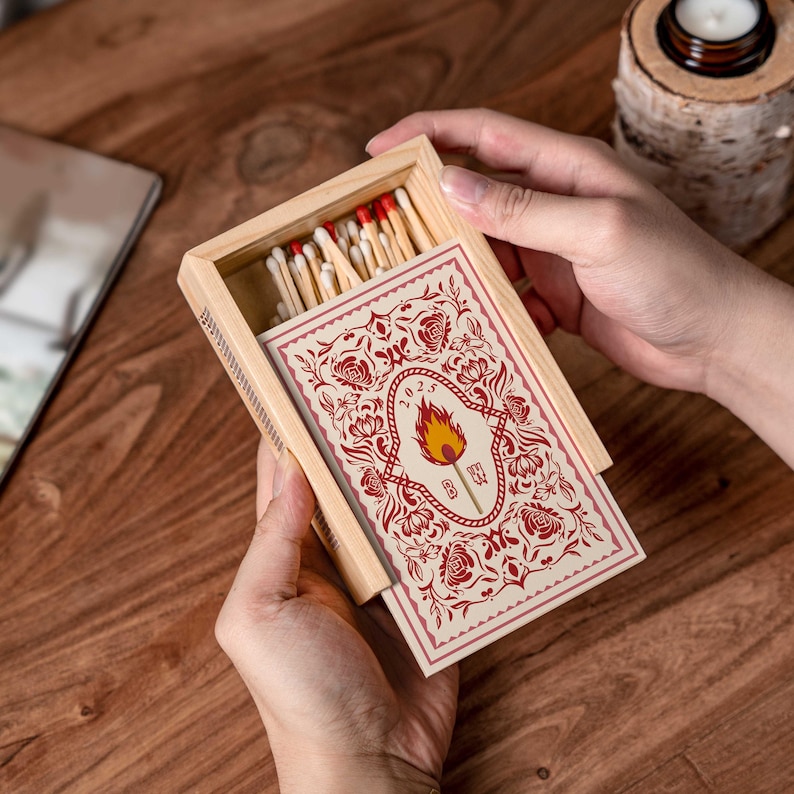 May include: A wooden matchbox with a decorative card featuring a flame design and the year 2023. The box is open, revealing red-tipped matches. The design includes floral elements in red and cream.