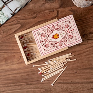 May include: A wooden matchbox with a decorative floral design in red and cream. The box is open, revealing matches with red tips. A pile of loose matches is scattered on the wooden surface.
