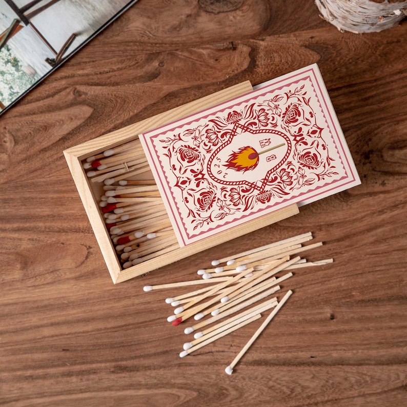 May include: A wooden matchbox with a decorative floral design in red and cream. The box is open, revealing matches with red tips. A pile of loose matches is scattered on the wooden surface.