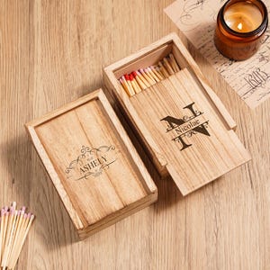 May include: A wooden matchbox engraved with the name "Nicolae" and the initial "N" on the lid, open to show matches with red tips. Another wooden box with the name "Ashely" is also visible. A lit candle in a glass jar is also present.
