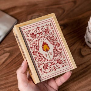 May include: A wooden matchbox with a decorative design. The matchbox features a cream-colored card with a red floral pattern and a central emblem of a torch with the year 2023. The box is held in a hand.