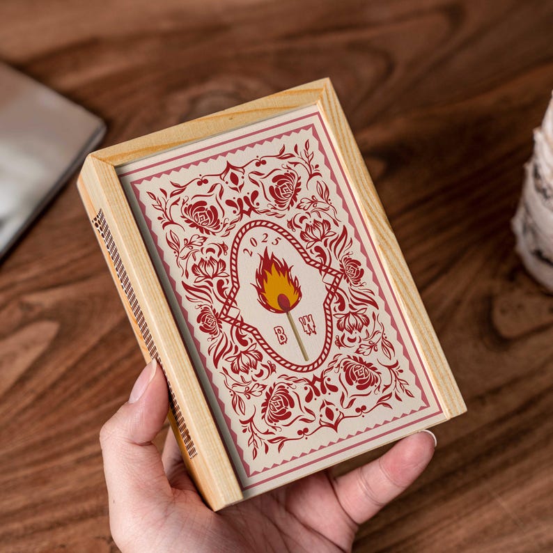 May include: A wooden matchbox with a decorative design. The matchbox features a cream-colored card with a red floral pattern and a central emblem of a torch with the year 2023. The box is held in a hand.