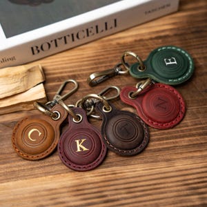 May include: A collection of leather keychains in various colors, including brown, burgundy, and green. Each keychain features a different letter in gold and a metal clasp. The keychains are arranged on a wooden surface.