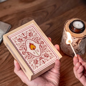 May include: A wooden matchbox with a decorative floral design in red and cream, featuring a central flame graphic and the year 2023. A lit match is held near a small candle in a glass jar, set on a wooden base. The scene is set on a wooden surface.