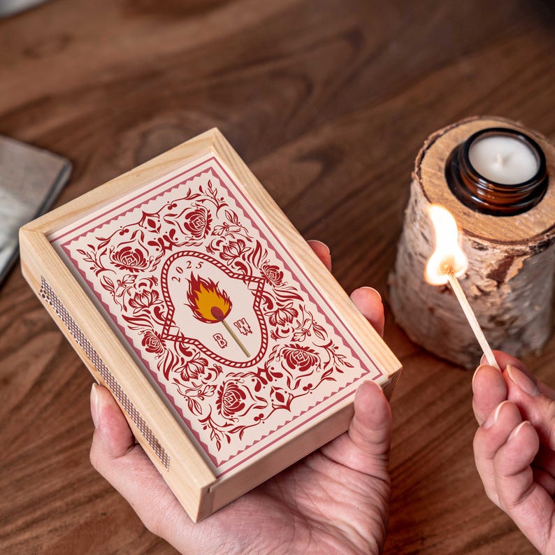 May include: A wooden matchbox with a decorative floral design in red and cream, featuring a central flame graphic and the year 2023. A lit match is held near a small candle in a glass jar, set on a wooden base. The scene is set on a wooden surface.