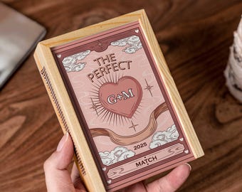 Puede incluir: Una caja de madera con un diseño rosa y blanco. El diseño presenta un corazón con las iniciales "G+M", las palabras "THE PERFECT" y el año "2025". La palabra "MATCH" también es visible. La caja se sostiene en una mano.