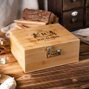 May include: A light brown wooden box with a combination lock. The lid is engraved with a crest, the name "RICHARD", and a date. The box is rectangular and sits on a wooden surface. The box is made of wood.