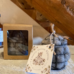 May include: A gray alpaca fiber bird nesting ball with a wire cage, a cardboard box with a clear window, and a tag that reads "Charity Driven Alpacas Bird Nesting Fiber". The items are on a white lace tablecloth.