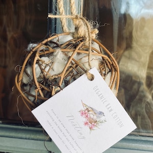 May include: A spherical bird nest made of twigs and filled with raw cotton, suspended by a jute rope. A tag with the text "ORGANIC RAW COTTON" and a bird illustration is attached. The tag also includes instructions for use.