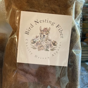 May include: A clear plastic bag filled with brown alpaca fiber for bird nesting. The bag has a white label with the text "Bird Nesting Fiber" and an illustration of an alpaca wearing flowers. The text "Charity Driven Alpacas" is also on the label.