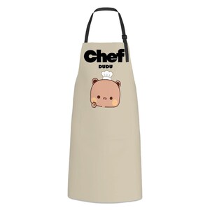 May include: A beige apron with black neck straps featuring the text Chef DUDU in black lettering above a graphic of a light brown cartoon bear wearing a white chef hat and making a hand gesture on a plain light beige background.