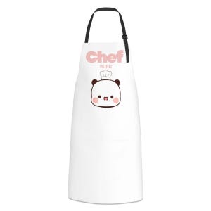 May include: A white apron featuring a black neck strap and a graphic print of a cartoon bear wearing a chef hat. The text Chef Bubu is printed in light pink above the bear illustration. The design is centered on the front of the white fabric.