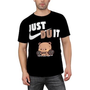 May include: Black t-shirt with the text "JUST DU IT" in white and brown. A cartoon bear lifting weights is featured below the text. The shirt is made of a soft material.
