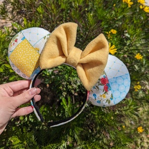 以下が含まれることがあります： 黄色と白の布製のミッキーマウスの耳に、黄色のリボンが付いている。耳はパッチワークのデザインで、花柄がプリントされている。