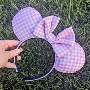 May include: Pink and white gingham-patterned mouse ears headband with a matching bow. The ears are round and padded, attached to a black headband. The bow is centered on the headband, adding a decorative touch. The headband is resting on green grass.