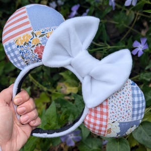 Peut inclure: Un serre-tête avec des oreilles de souris en tissu patchwork aux motifs floraux, vichy et rayés. Un grand nœud blanc est placé entre les oreilles. Le serre-tête est noir et blanc.