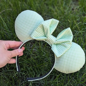 May include: A light green gingham-patterned headband with two round ears and a matching bow. The ears and bow are made of fabric with a white and light green checkered design. The headband is black with a light green trim.