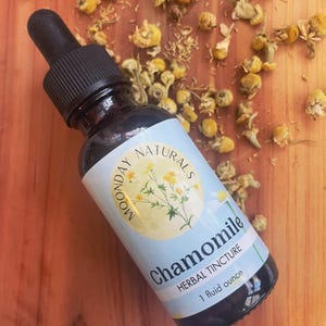 May include: A glass bottle with a dropper top filled with a brown liquid. The label is blue and white with the text "Moonday Naturals Chamomile Herbal Tincture 1 fluid ounce".