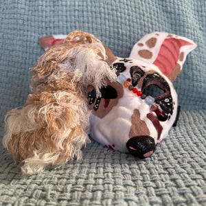 May include: A handmade dog mask with a white base, brown spots, and red accents on the ears. The mask features black and red details around the eyes and nose, with a beaded necklace. A fluffy, light brown and white fur piece is attached to the side.