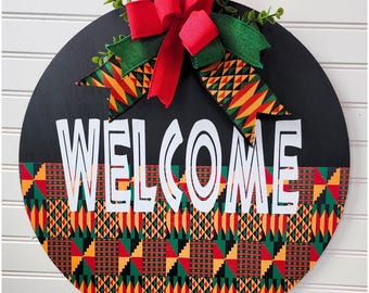 May include: A round wooden door hanger with a black background and a welcome sign in white. The bottom half of the door hanger is decorated with a colorful African print pattern in red, yellow, green, and black. A red bow with green leaves is attached to the top of the door hanger.