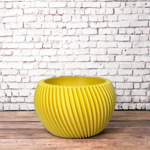 Könnte beinhalten: Ein leuchtend gelber Topf mit geripptem Design. Der Topf steht auf einer Holzoberfläche vor einer weißen Backsteinwand.