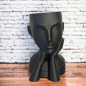 Könnte beinhalten: Ein schwarzer, stilisierter, menschlicher Büsten-Pflanztopf mit einem Frauenkopf. Der Topf steht auf einer Holzoberfläche.