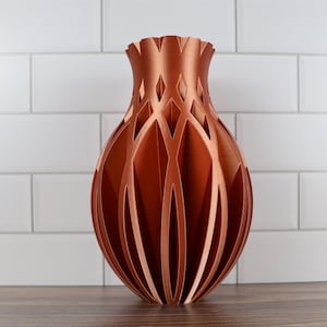Peut inclure: Un vase décoratif de couleur cuivre avec un design ajouré unique. Le vase a un dessus évasé avec un bord festonné et une série d'ouvertures verticales et incurvées. Le vase est en matériau métallique et repose sur une surface en bois.