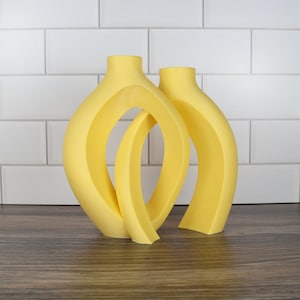 Könnte beinhalten: Zwei gelbe Keramikvasen mit abstraktem Design. Die Vasen sind übereinander gestapelt und haben einen modernen, minimalistischen Stil.
