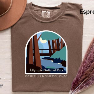 Puede incluir: Camiseta marrón espresso con un gráfico de una escena de bosque con un puente y un río. El gráfico está en un círculo blanco con el texto "Olympic National Park" y "Protect Our National Parks".