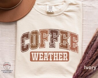 Könnte beinhalten: Elfenbeinfarbenes T-Shirt mit dem Text "COFFEE WEATHER" in einem verwitterten, braunen Schriftzug. Ein brauner Hut und Trockenblumen sind im Hintergrund. Das Shirt ist ein Comfort Colors XL.
