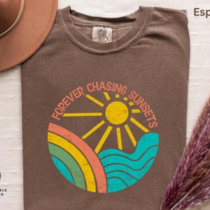 May include: Espresso-coloured t-shirt with a circular graphic reading "Forever Chasing Sunsets". The design features a yellow sun, rainbow, and blue waves. A brown hat and dried flowers are in the background.