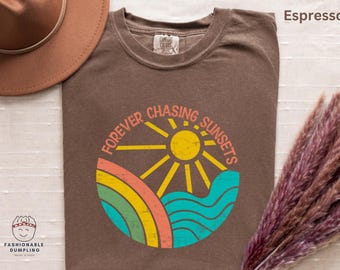 May include: Espresso-colored t-shirt with a circular graphic that reads "Forever Chasing Sunsets." The design features a yellow sun, rainbow, and blue waves. A brown hat and dried flowers are in the background.