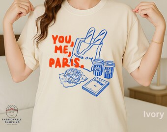 Puede incluir: Camiseta de color marfil con un diseño gráfico. El diseño incluye el texto "YOU, ME, PARIS" en rojo, una baguette en una bolsa de mano, dos tazas de café y un pastel. La camiseta es de estilo casual.