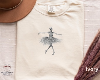 Può includere: T-shirt color avorio con una grafica di ballerina scheletrica in grigio e nero. La ballerina indossa un tutù. La maglietta ha girocollo e maniche corte. Il testo "Ivory" è stampato in basso a destra.