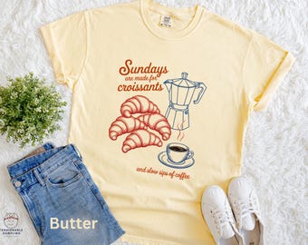 May include: A pale yellow t-shirt featuring a graphic of croissants, a coffee pot, and a coffee cup. The text reads "Sundays are made for croissants and slow sips of coffee." The shirt is styled with blue jeans and white trainers.