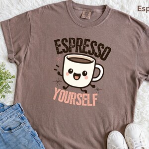 Op de afbeelding: Espresso-kleurig T-shirt met de tekst "Espresso Yourself" en een cartoon koffiekopje. Het shirt is opgevouwen, met een blauwe jeans, witte sneakers en een kleine plant op de achtergrond.