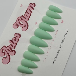 May include: A set of mint green press-on nails in various sizes, arranged on a white card. The card features the text "Aries Glam" in a pink, retro font, along with Instagram contact information. The nails have a matte finish and a pointed shape.